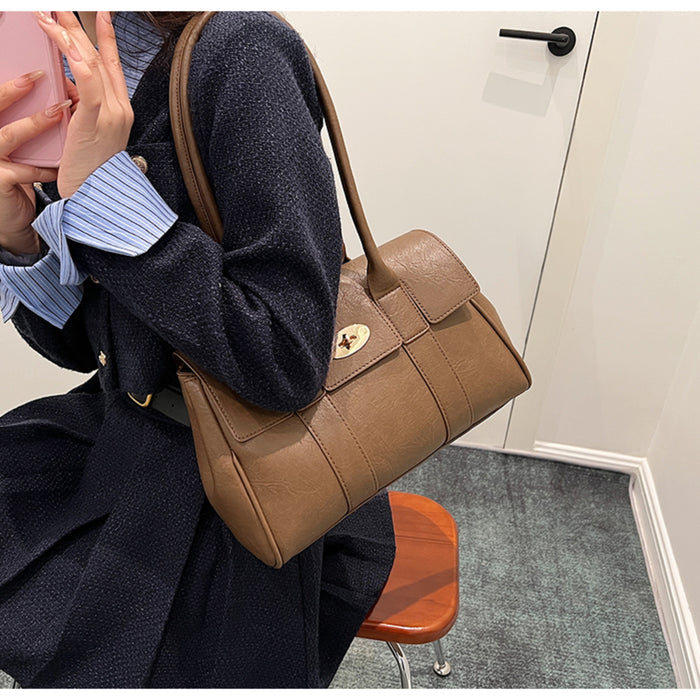 Stylish woman wearing a navy coat holding a brown leather shoulder bag with a gold clasp, seated indoors with a modern minimal backdrop