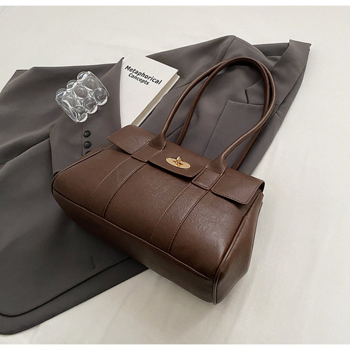 Dark brown leather shoulder bag with turn-lock closure placed on a grey blazer next to a book and clear ring accessory, styled flatlay for fashion presentation