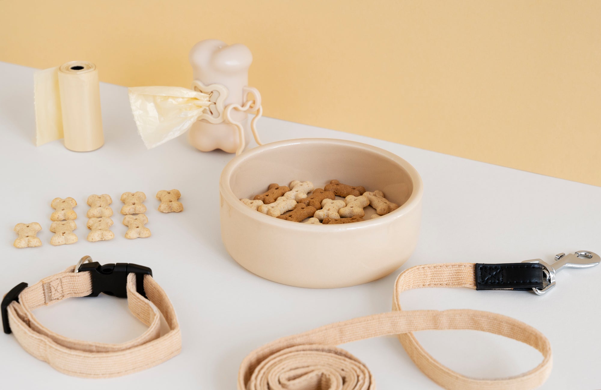 Close-up of dog accessories including a beige collar, leash, dog bone-shaped treats, and waste bag dispenser on a white surface with a soft yellow background.